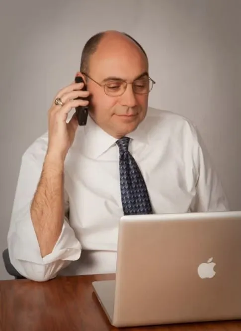Man in white shirt on a phone call, looking at laptop.