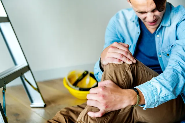Man clutching knee after falling near ladder, yellow hard hat on floor.