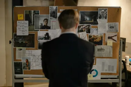 Person in suit facing a corkboard covered with photos, notes, and charts, possibly investigating something.