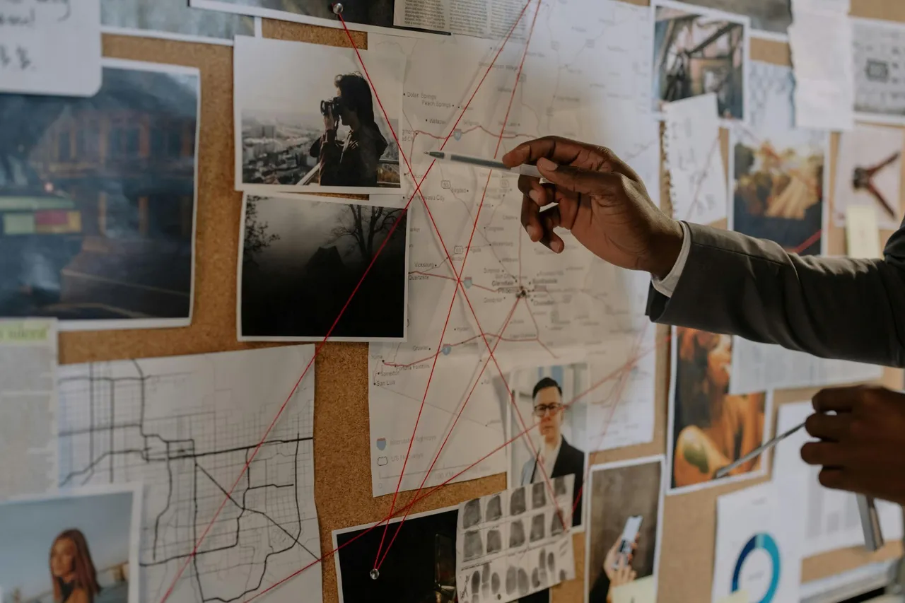 Person pointing at photos and map on a corkboard, connecting them with red string, detective board.